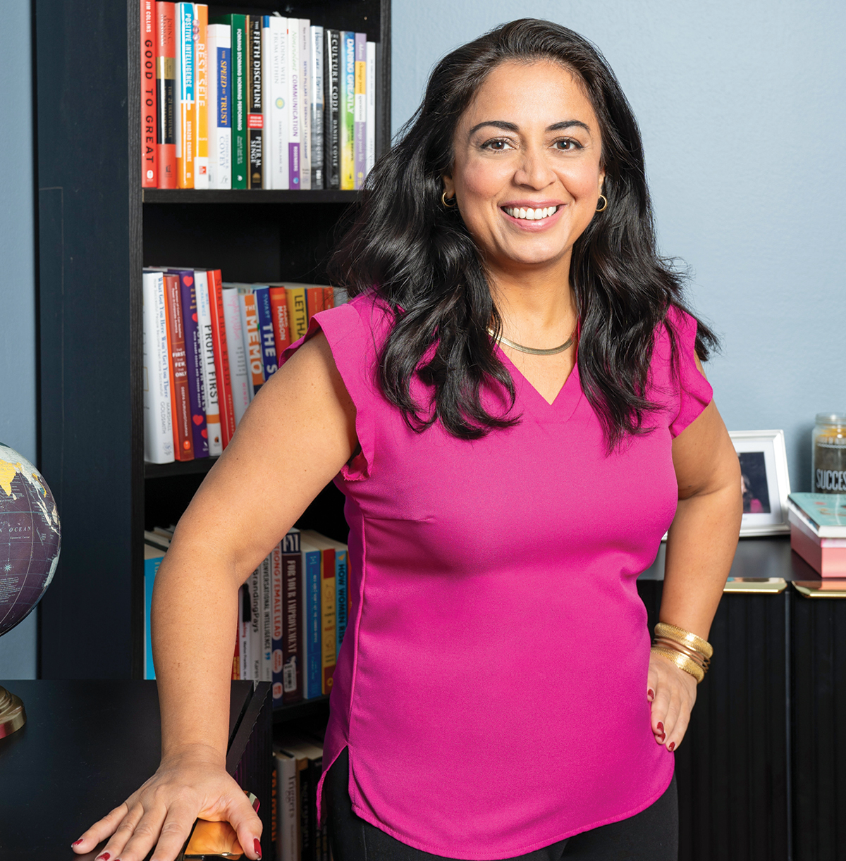 A woman with black hair wearing a pink shirt standing in front of a bookcase