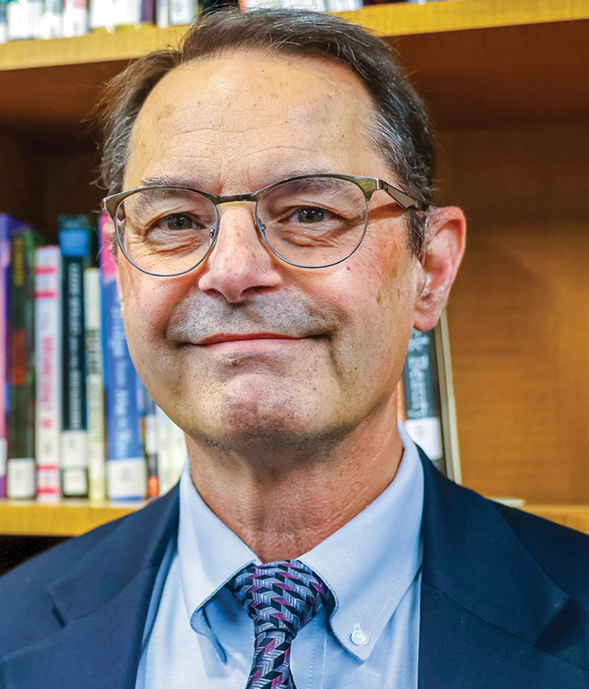 smiling headshot of brunette man wearing glasses and suit in front of bookcase